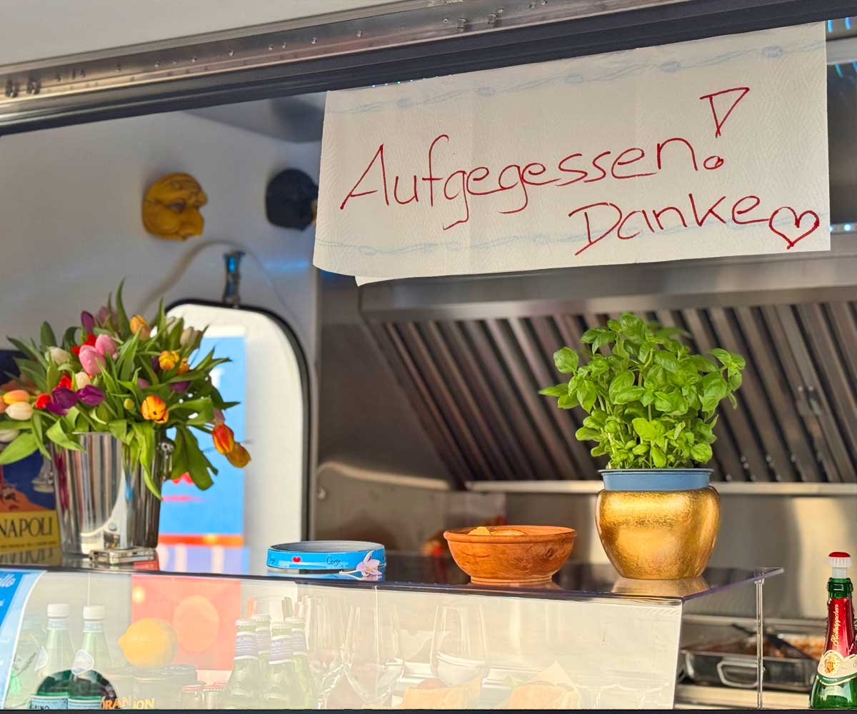 Ein Zettel auf dem die Worte aufgegessen, Danke stehen hängt an der Durchreiche eines Foodtrucks. Auf dem Tresen stehen bunte Blumen und ein Basilikum-Topf.