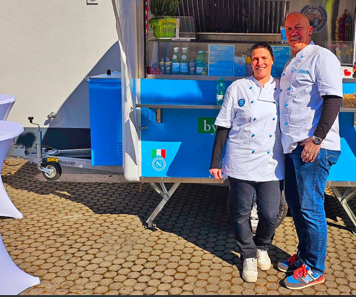 Ein Mann und eine Frau dicht nebeneinander stehend, in weiß-blauen Kochjacken lächend vor einem blauen Foodtruck.