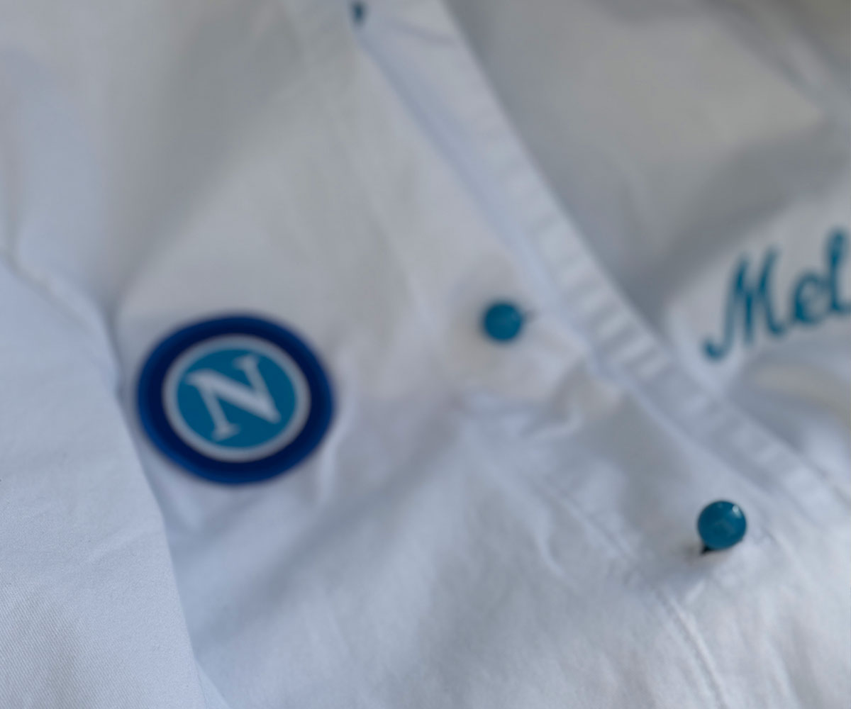 Eine weiße Kochjacke auf der ein blaues N, blaue runde Knöpfe und der name Mel eingestickt ist.