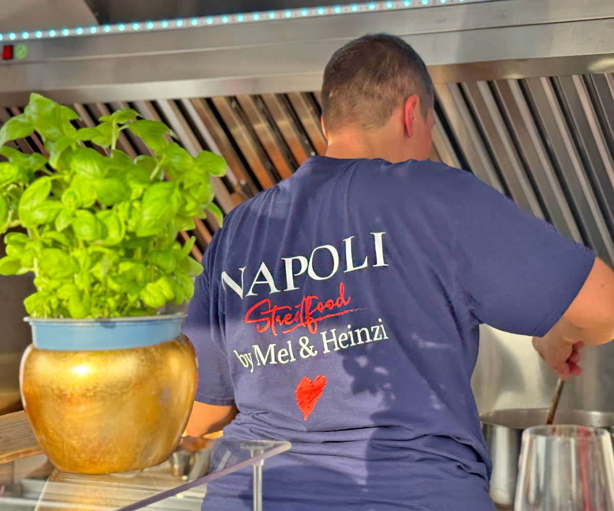 Foodtruck-Köchin in Bamberg bereitet frisches Cuzzetiello im Napoli Streetfood-Truck zu. Man sieht sie von hinten während sie mit einem Kochlöffel im Topf rührt, auf ihrem blauen T-Shirt ist das Logo und "by Mel und Heinzi" zu lesen.