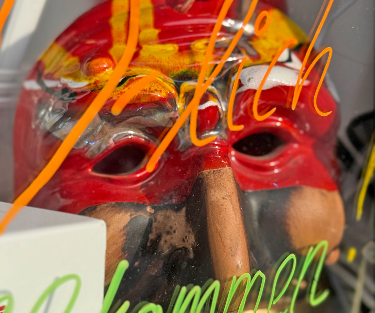 Neapolitanische rote Maske liegt hinter einer Glasscheibe die mit bunten Farben beschrieben ist. Ein Herz und Buchstaben sind zu sehen.