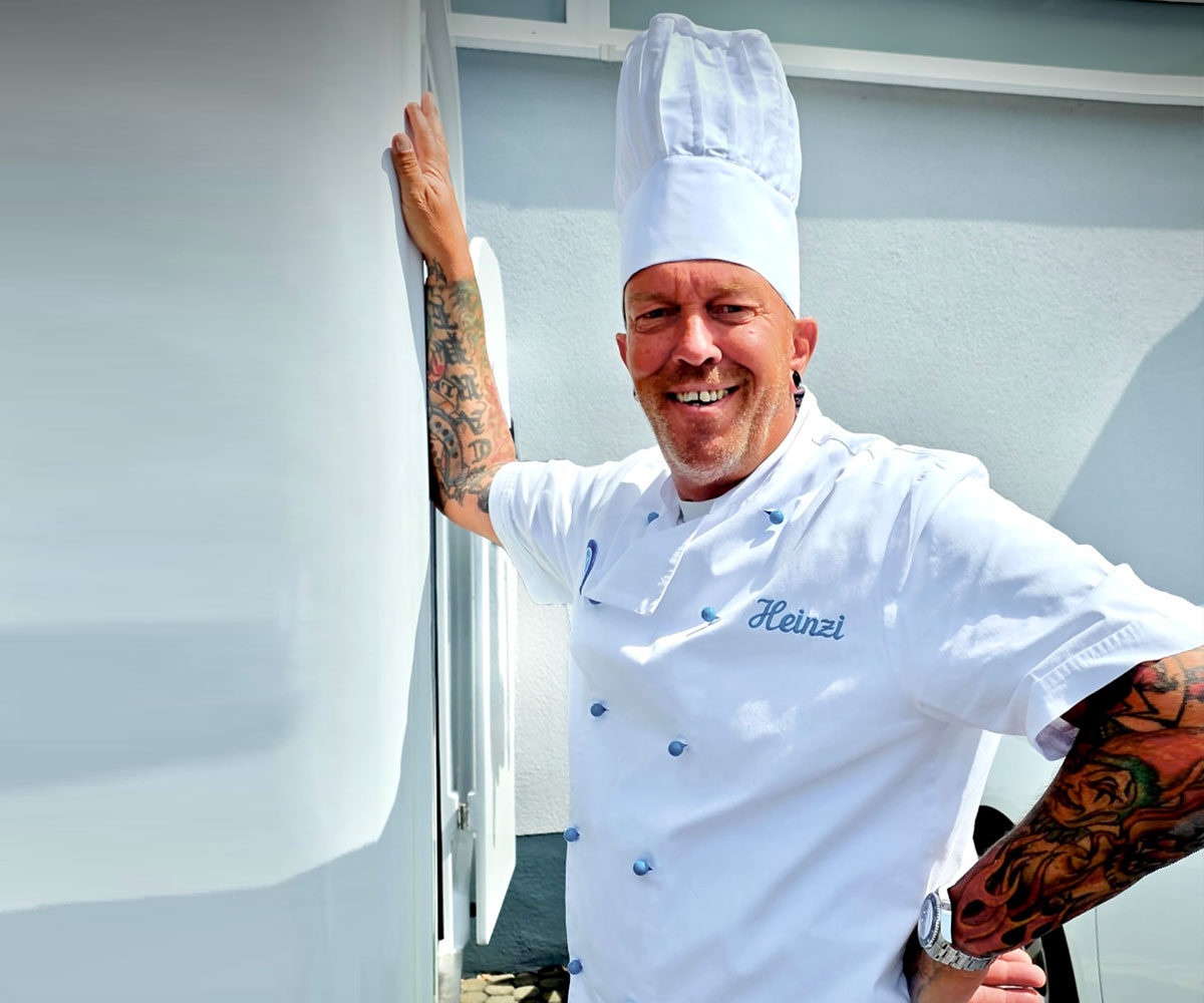 Lächelnder Mann trägt weiße Kochjacke und eine hohe Kochmütze und lehnt mit tätowierten Unterarmen an einem Foodtruck.