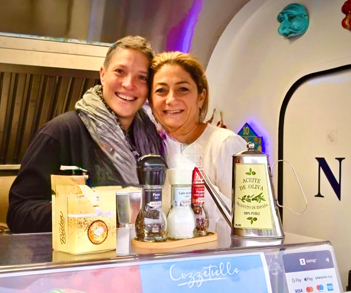 Zwei Frauen stehen lächelnd, freudig, Wange an Wange nebeneinander hinter dem Tresen eines Foodtrucks.