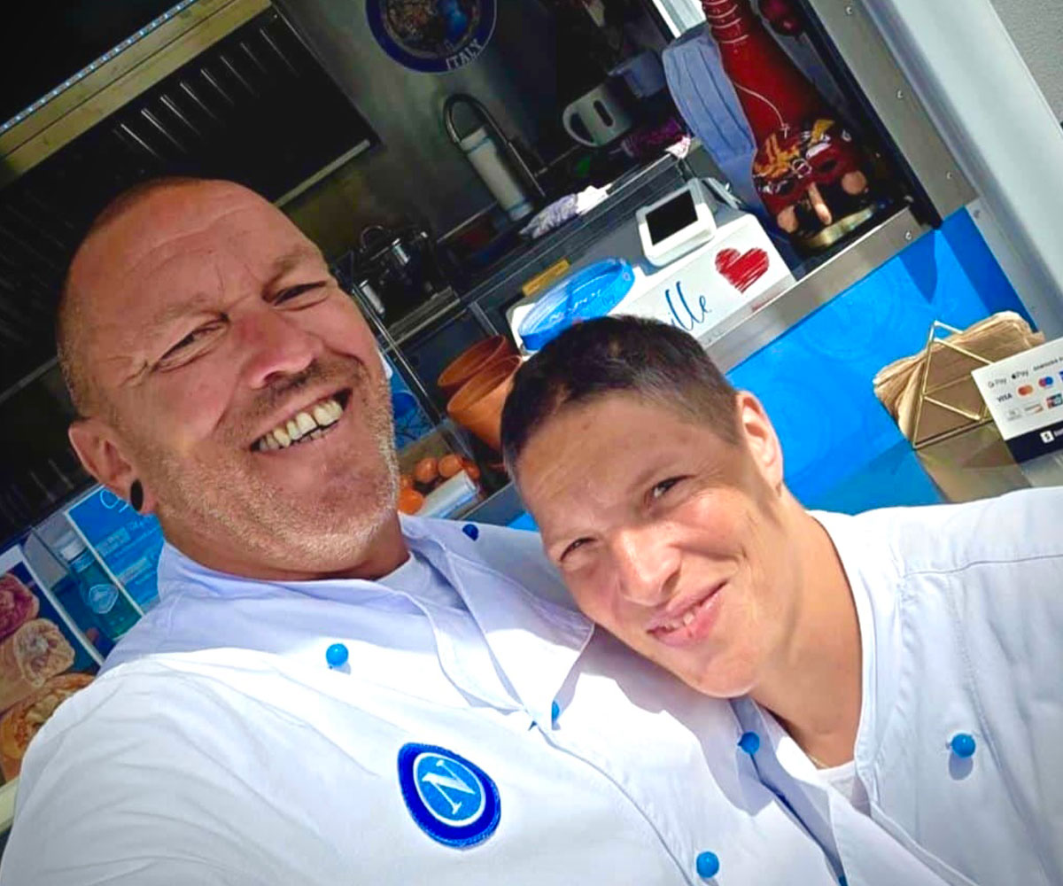 Großer Mann hält kleinere, kurzhaarige Frau im Arm. Beide tragen weiße Kochjacken mit blauen Knöpfen und stehen lächelnd vor einem Foodtruck.