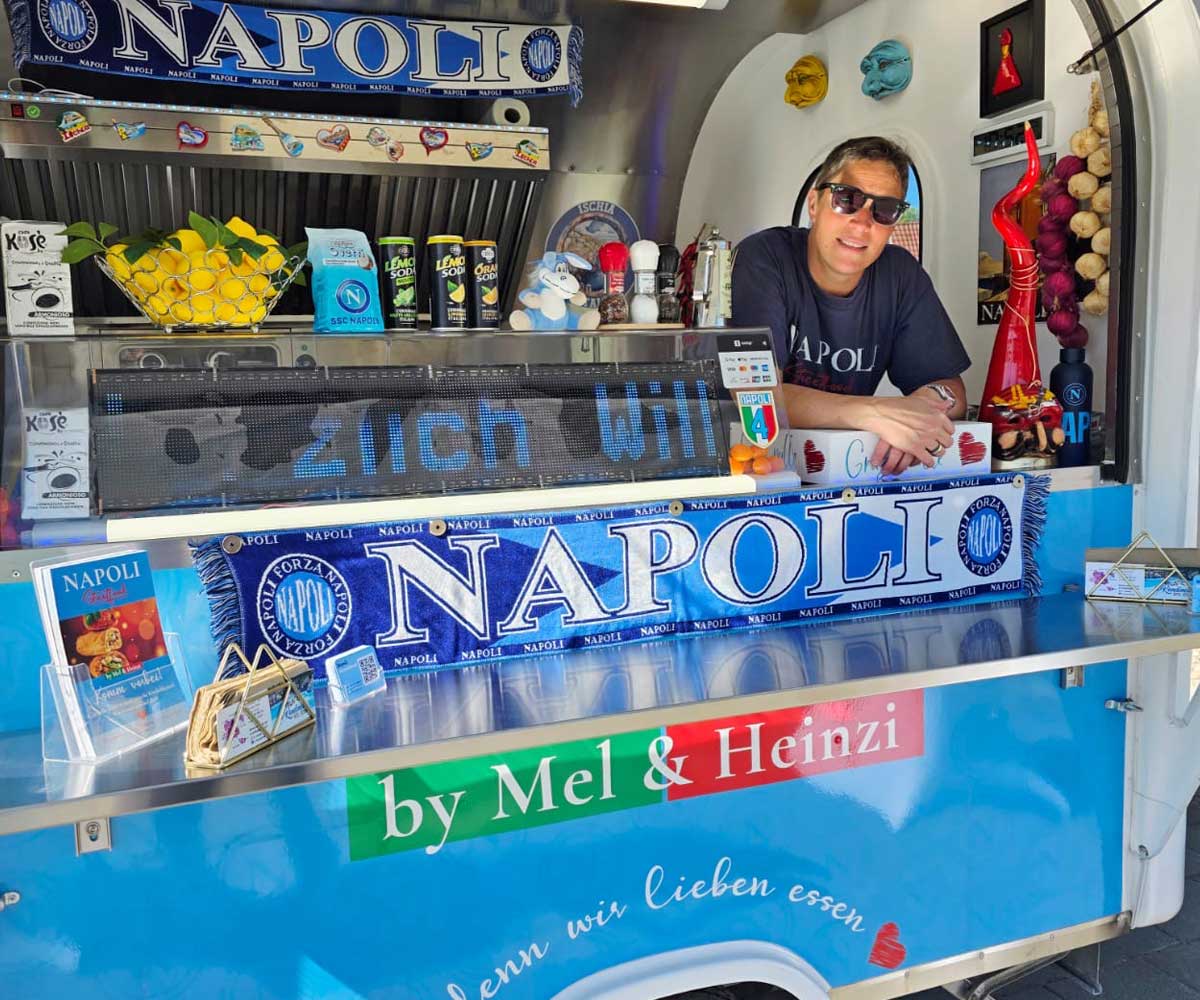 Frau mit Sonnenbrille steht in einem Foodtruck und leht leger auf der Kasse am Tresen. Der Foodtruck ist sichtlich in blau und weiß geschmückt.