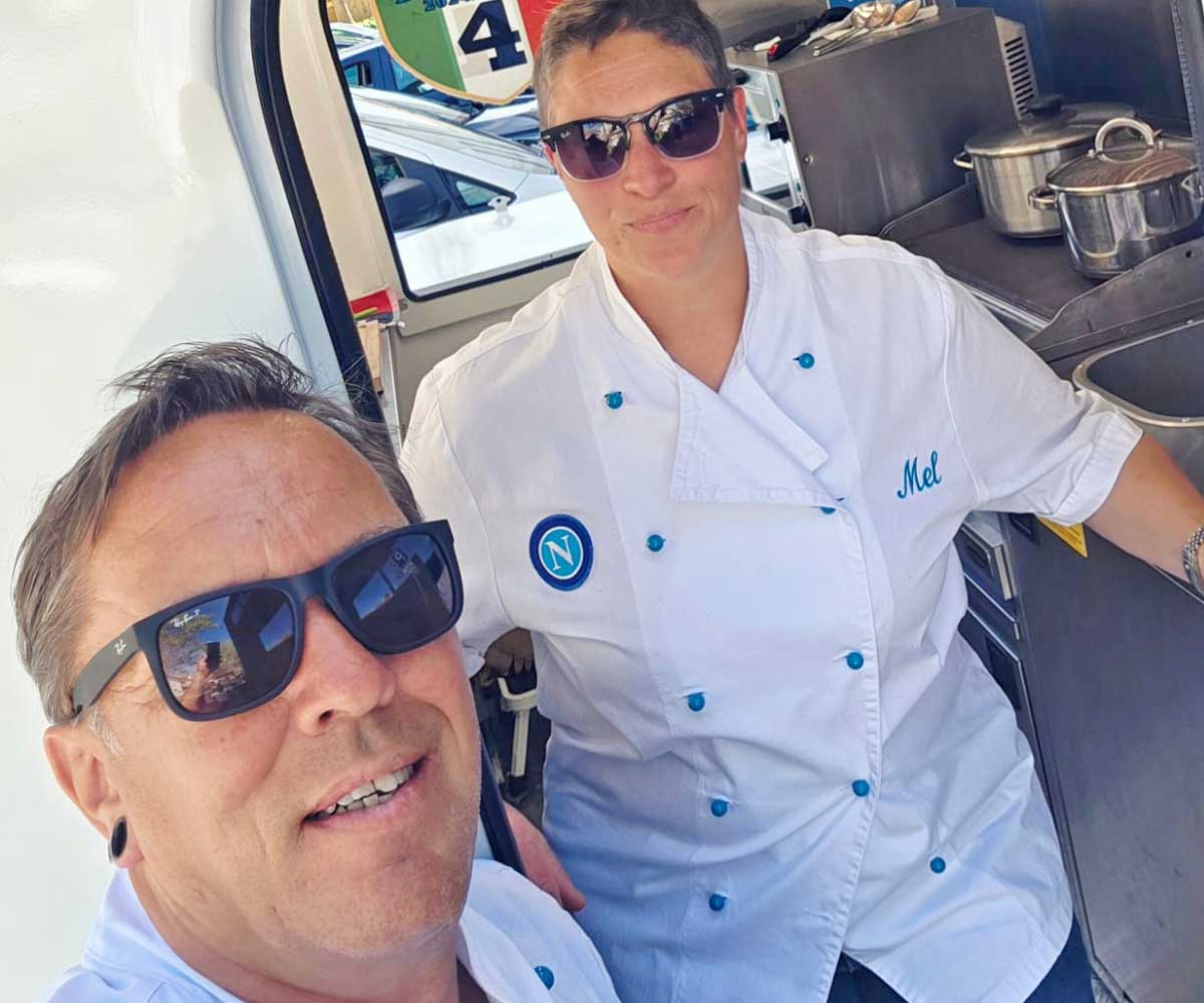 Mann und Frau tragen weiße Kochjacken, die blaue Knöpfe haben und stehen in der Tür eines Foodtrucks.