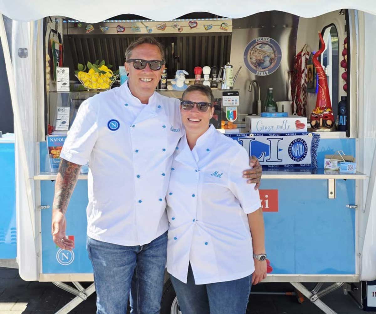 Mann und Frau tragen weiße Kochjacken, die blaue Knöpfe haben umarmen sich und stehen vor einem Foodtruck .